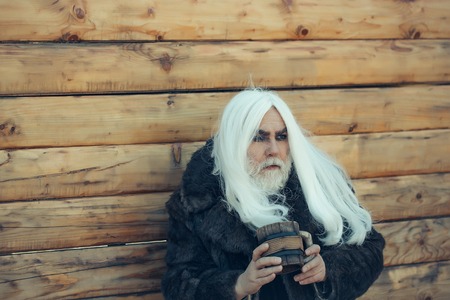 Druid old man with long grey hair and beard on serious face with wooden mug in hands outdoor on wood backgroundの写真素材