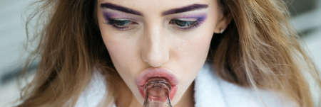 Young woman with pretty face professional vogue makeup lilac eye shadow healthy skin drinking from glass bottle closeup panoramic imageの写真素材