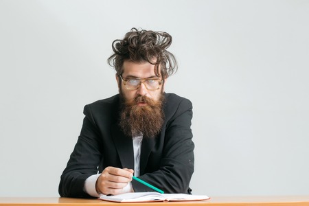 young handsome bearded crazy emotional man scientist or professor with long beard and teacher glasses with pencil and book or notepaper sitting at table on grey backgroundの写真素材