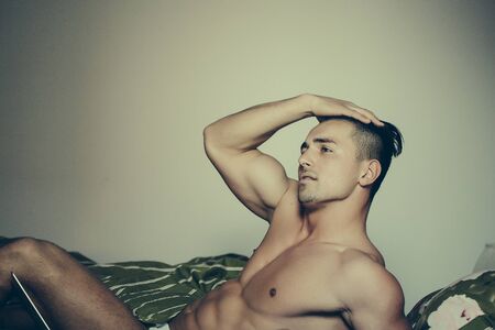 Handsome young man with athletic muscular body and bare torso laying in bed in morning with laptop