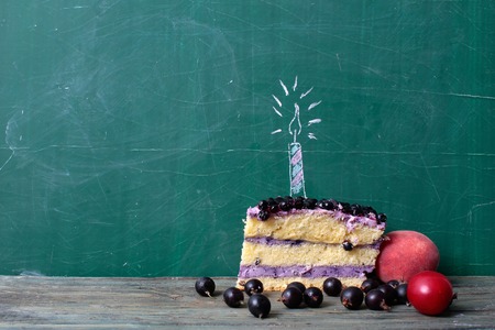 piece of sweet blueberry berry cake with cream and painted candle for birthday party on green school blackboard near currant plum and peach on tableの写真素材
