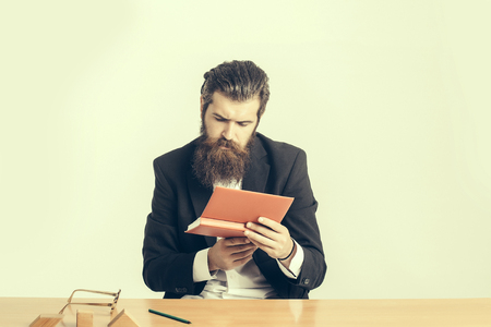 young handsome bearded man scientist or professor with long beard and teacher glasses with book or notepaper sitting at table with wooden cubes on light backgroundの写真素材