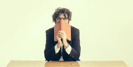 young handsome bearded crazy emotional man scientist or professor with long beard and teacher glasses with pencil and red book or notepaper sitting at table isolated on white backgroundの写真素材