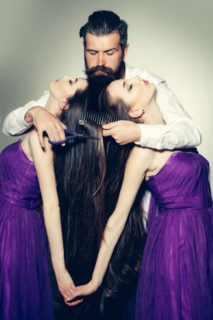 Bearded handsome man with scissors and brush in white shirt with two young pretty women in violet dresses with long hair in studio on grey backgroundの写真素材
