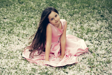 Beautiful little girl in pink dress with long brunette hair and thoughtful face sitting on green grass covered with spring flower blossom petals outdoorの写真素材