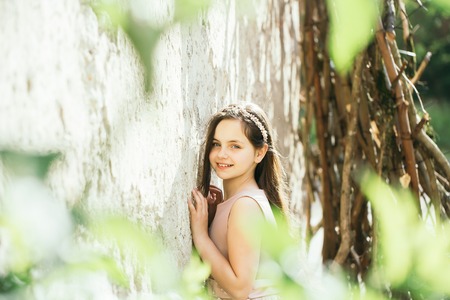 Cute little girl with smiling face in pink dress and wreath standing near white wall in green leaves sunny day outdoorの写真素材