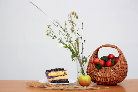 Wicker basket full of red plums bunch of wild flowers in glass vase piece of tasty cake on plate and green apple for lunch on brown wooden school desk on white backgroundの写真素材