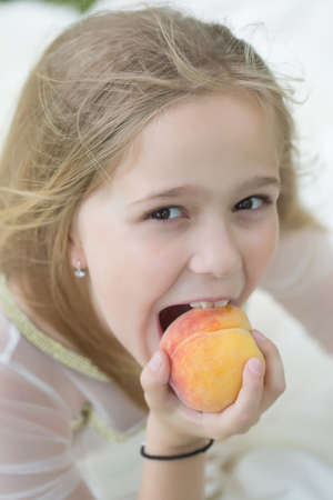 Pretty blond girl eats fresh sweet delicious orange peach fruit in white dressの写真素材