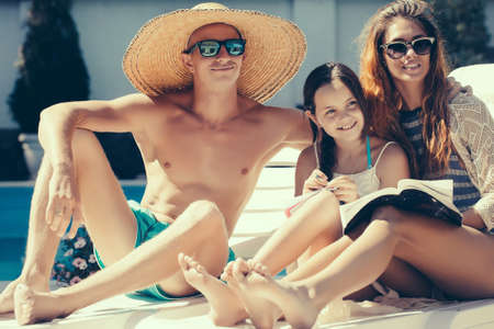 young happy family couple of pretty woman in glasses near muscular man in straw hat and small girl reading book at swimming pool with blue water sunny summer vacationの写真素材