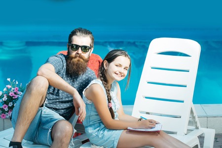 young handsome bearded man with long beard in sun glasses on face on beach chair and small happy girl with pen and notebook at swimming pool with blue water sunny summer vacationの写真素材