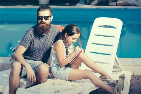young handsome bearded man with long beard in sun glasses on face on beach chair and small happy girl with pen and notebook at swimming pool with blue water sunny summer vacationの写真素材