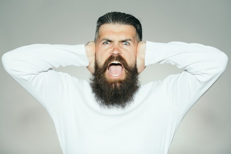handsome young man with long beard and moustache on emotional face with hands on ears shouting on...