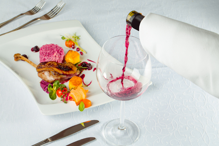 Confit duck or goose leg crisp delicious with colorful garnish on blackberry sauce served in restaurant with cutlery and red wine on white backgroundの写真素材