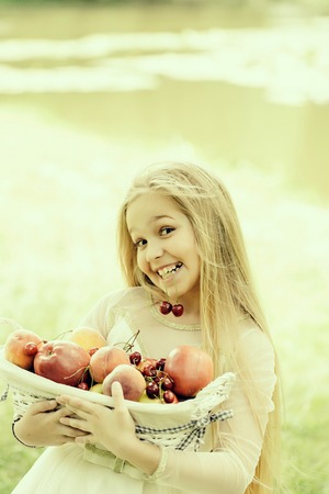 small girl kid with long blonde hair and pretty smiling happy face in prom princess white dress standing sunny day outdoor near water with fruit basket of red apples peach and cherryの写真素材