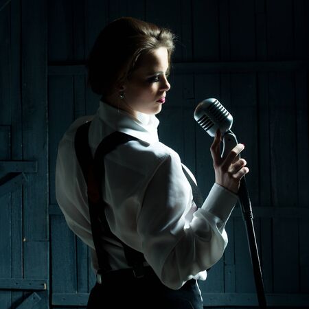 young pretty woman with sexy stylish cloth singing into silver studio microphone in retro braces and pants in darknessの写真素材