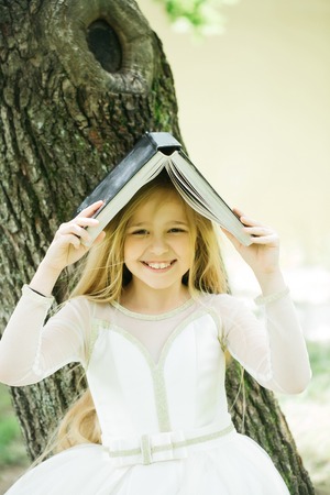small girl kid with long blonde hair and pretty smiling happy face in prom princess white dress standing sunny day outdoor near water with bookの写真素材