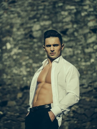 Man bare-chested young handsome sensual model in white shirt gaped open poses with hands in black trouser pockets outside on masonry backgroundの写真素材