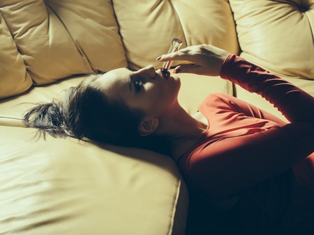 Pretty girl paints her lips red on the couch. Sexy woman with red lips and in red dressの写真素材