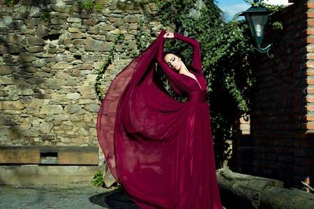 Young beautiful woman with brunette hair in fluttering red dress outdoor near brick stony wallの写真素材