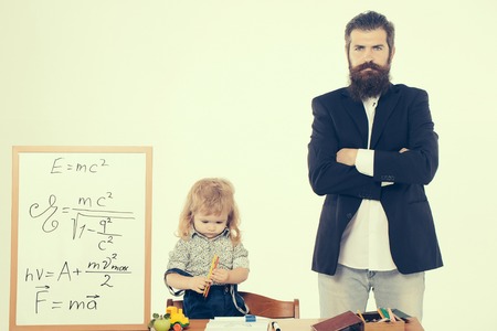 Cute boy small child in shirt sitting at school desk with colored pencils near blackboard with formulas and standing bearded professor man isolated on white backgroundの写真素材