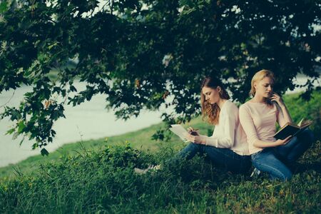 Two young girls best friends sit on green grass read and write in summer parkの写真素材