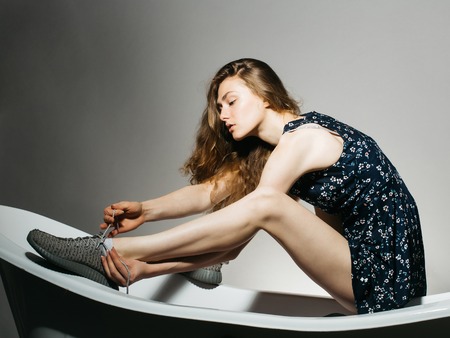 Young woman with pretty face brown hair long sexy beautiful legs dressed in dark dress sitting on bath and tieing shoelaces on her sport shoes studio on gray backgroundの写真素材