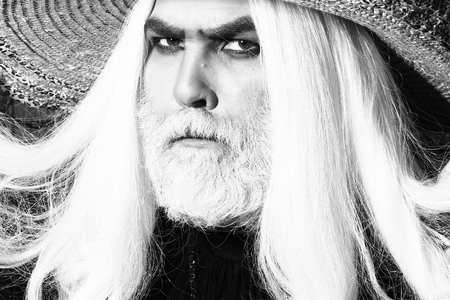 Bearded senior man in long white wig and straw round hat on wooden background, black and white