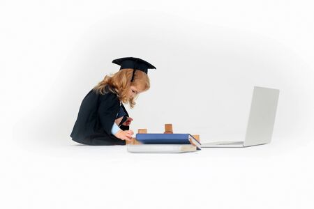 Small boy child with long blond hair in blue shirt black graduation gown and cap sitting and playing with wooden blocks near notebook and diaries isolated on white backgroundの写真素材