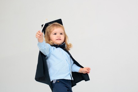 Little boy child with smiling face long blond hair in blue shirt black graduation squared cap and mantle holding colored pencil in hand isolated on white backgroundの写真素材