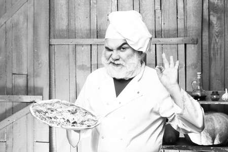 Senior bearded chef with pizza in white uniform with winking face and hat on wooden backgroundの写真素材