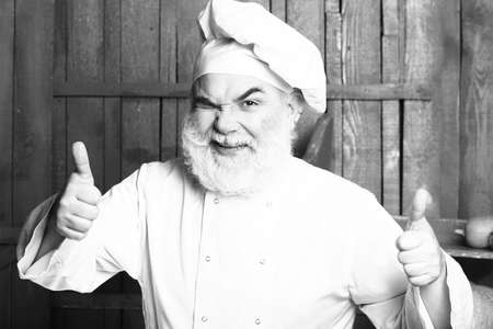 Bearded man cook with winking face in chef hat with thumb up in studio on wooden background, black and whiteの写真素材