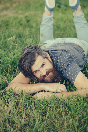 handsome sexy bearded young man hipster with long beard and mustache on happy smiling face relax and laying on green grass field outdoor on natural backgroundの写真素材
