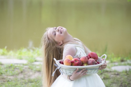 small girl kid with long blonde hair and pretty smiling happy face in prom princess white dress standing sunny day outdoor near water with fruit basket of red apples peach and cherryの写真素材