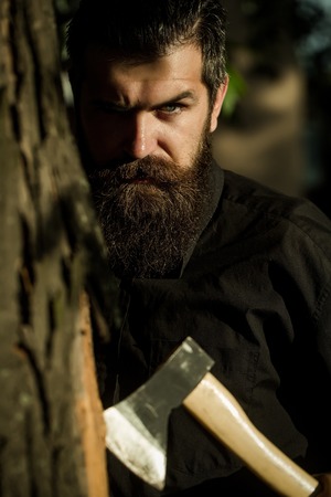 handsome bearded young man hipster with long beard and mustache has stylish hair on serious face sunny day outdoor holding sharp axe or ax on green natural backgroundの写真素材