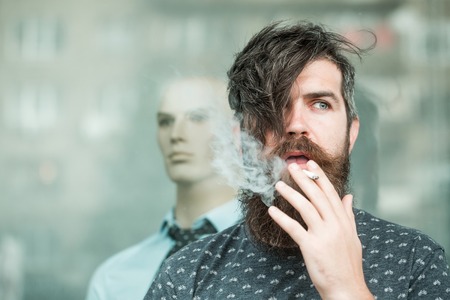 handsome bearded young man hipster with long beard and mustache has stylish hair smoking cigarette near shop glass showcase with dummy in shirt and tieの写真素材