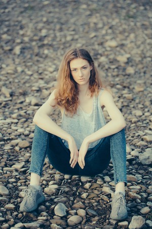 Young pretty woman in blue jeans and sleeveless vest sits on pebble beach on summer dayの写真素材