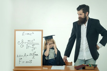 Cute child little boy in black academic gown squared hat and glasses at desk with pencils in box near school chalkboard and teacher man with beard isolated on white backgroundの写真素材