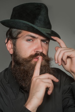 handsome bearded young man hipster or gentleman with long beard and mustache in stylish black retro hat on serious face in studio on grey background, portraitの写真素材