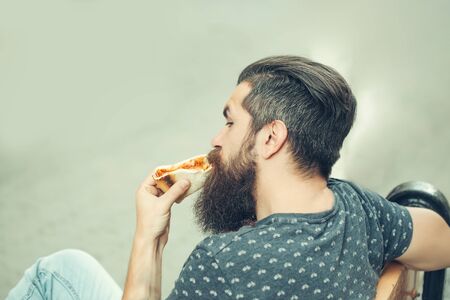 handsome sexy bearded young man hipster with long beard and mustache on serious hairy face eating pizza sitting on street bench outdoorの写真素材