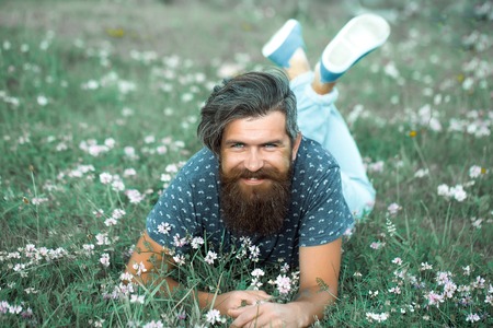 handsome sexy bearded young man hipster with long beard and mustache on happy smiling face relax and laying on green grass field with wild flowers outdoor on natural backgroundの写真素材