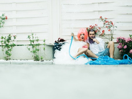 Young girl with pretty smiling face pink hair in beautiful dress holding blue hose sitting on ground with handsome bearded man with lollipop in his hand near flowers on white background copyspaceの写真素材