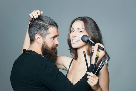 young couple of sexy woman with pretty smiling face and long brunette hair in black bra on body and handsome bearded man visagiste with fashionable makeup brush in studio on grey backgroundの写真素材