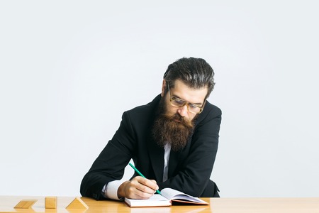 young handsome bearded man scientist or professor with long beard and teacher glasses with book or notepaper sitting at table with wooden cubes isolated on white backgroundの写真素材