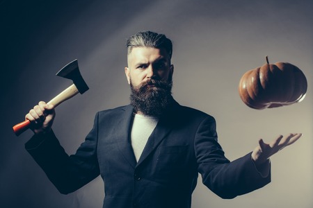 Handsome young man with long beard and moustache in black jacket holding halloween pumpkin and axe in studio on grey backgroundの写真素材