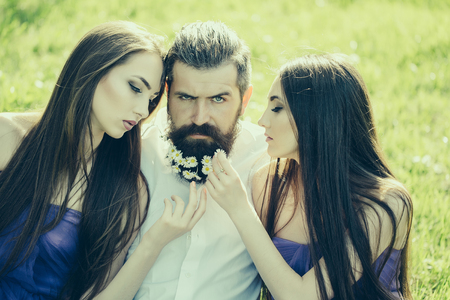 handsome man in white shirt with dandelion flowers in beard with two young pretty women in violet dresses on green grass sunny day outdoorの写真素材