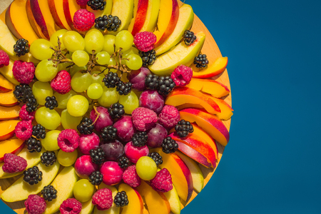 Fruit and berry platter with ripe fresh sweet vibrant healthy organic red raspberry peach blackberry green grapes and on round plate on blue background copy spaceの写真素材