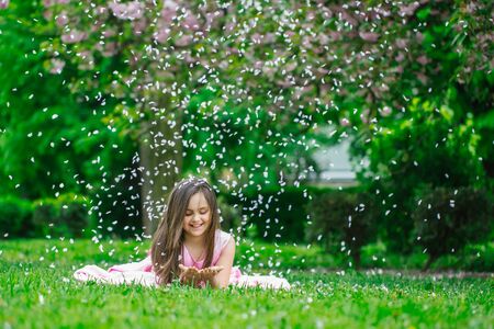 Beautiful little girl in pink dress with long brunette hair and smiling face lying on green grass in spring flower blossom petals outdoorの写真素材