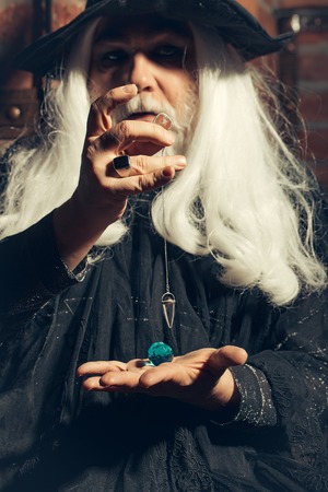 Old man wizard with long gray hair beard in black costume and hat for Halloween holding blue gem stone and silver pendant for hypnosis on wooden backgroundの写真素材
