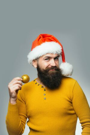 handsome bearded man with long lush beard and moustache on happy smiling face decoration ball in yellow shirt and christmas hat on grey backgroundの写真素材