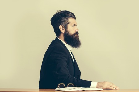 young handsome bearded man scientist or professor with long beard and teacher glasses with book or notepaper sitting at table on light backgroundの写真素材